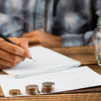 Pessoa anotando valores ao lado de moedas e comprovantes