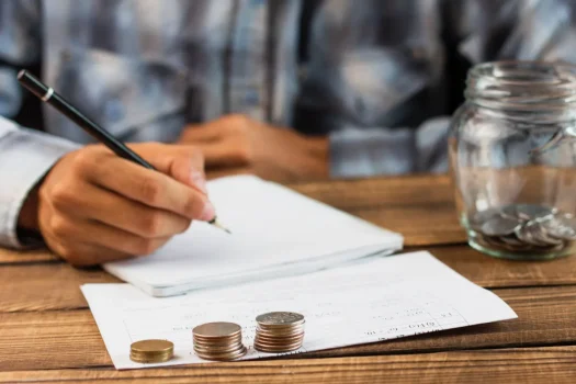 Pessoa anotando valores ao lado de moedas e comprovantes