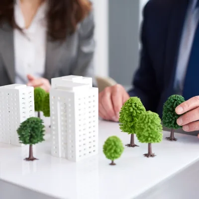 Duas pessoas de terno em uma mesa, observando maquetes de prédios e árvores, representando o planejamento e a discussão sobre erros na gestão condominial e melhorias urbanas.