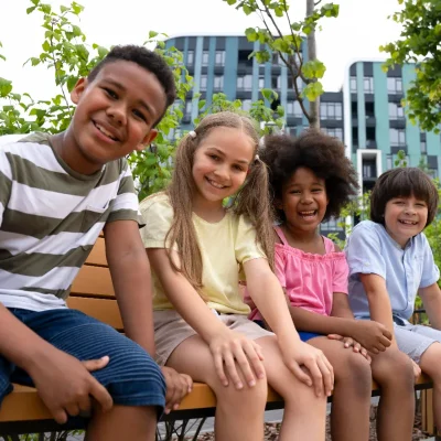 Quatro crianças de diferentes etnias, sorrindo e sentadas em um banco de madeira em uma área verde de lazer com prédios ao fundo, simbolizando a convivência e a necessidade de segurança para crianças no condomínio.