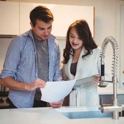 Casal em uma cozinha moderna olhando a conta de água do condomínio e um tablet sobre a pia, próximo a uma torneira metálica. A imagem aborda a preocupação dos moradores com os custos hídricos e a necessidade de controle e transparência na gestão da cobrança, que pode ser coletiva ou individualizada e deve seguir as novas regras do STJ. Também sugere o uso de tecnologia para redução de desperdícios e economia de água.