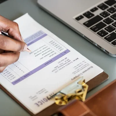 Close-up de uma mão preenchendo uma planilha ou folha de pagamento com caneta sobre uma prancheta, com um laptop ao lado, simbolizando a elaboração e o cálculo da folha de pagamento do condomínio, incluindo salários, encargos e descontos.