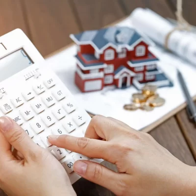Uma pessoa utilizando uma calculadora para determinar o valor do aluguel, enquanto ao fundo estão moedas, um modelo de casa e documentos relacionados ao imóvel.