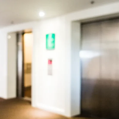 Imagem desfocada (bokeh) de um hall de entrada de edifício com portas de dois elevadores em condomínio, simbolizando a acessibilidade e a segurança no transporte vertical. A gestão desses equipamentos exige manutenção regular e o cumprimento de normas técnicas como a NBR 12892 da ABNT.