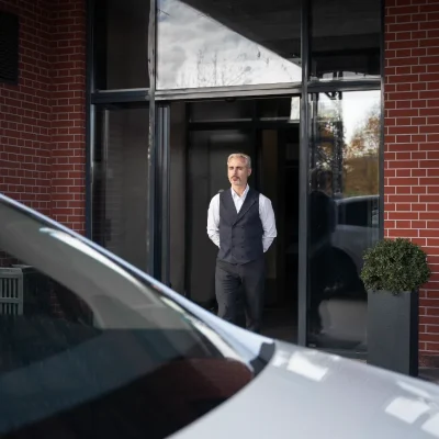 Porteiro com uniforme social (colete e camisa social) parado na porta de um prédio. A imagem representa um dos tipos de portaria em condomínios: a portaria tradicional (presencial). Os tipos de portaria em condomínios incluem os modelos tradicional, remoto, híbrido e autônomo, cada um com diferentes impactos na segurança, controle de acesso e custos. A portaria é uma decisão estratégica na gestão condominial.