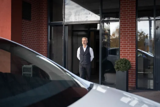 Porteiro com uniforme social (colete e camisa social) parado na porta de um prédio. A imagem representa um dos tipos de portaria em condomínios: a portaria tradicional (presencial). Os tipos de portaria em condomínios incluem os modelos tradicional, remoto, híbrido e autônomo, cada um com diferentes impactos na segurança, controle de acesso e custos. A portaria é uma decisão estratégica na gestão condominial.