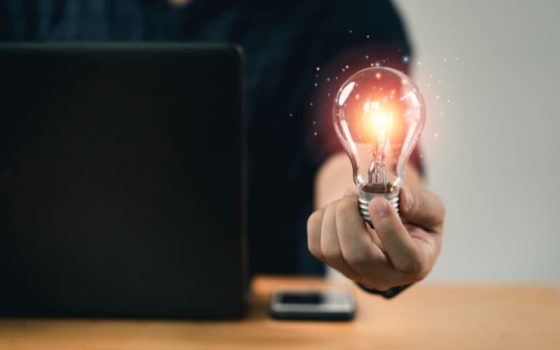 Imagem de um homem branco segurando uma lâmpada em frente a um notebook e um celular, representando inovações para 2024.