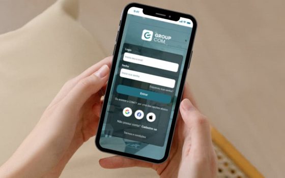 Imagem de duas mãos segurando um celular com a tela de login do GroupCOM.