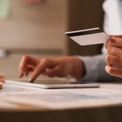 Pessoa segurando um cartão de crédito ao lado de um tablet e documentos, em imagem sobre se condomínio pode ter cartão de crédito.